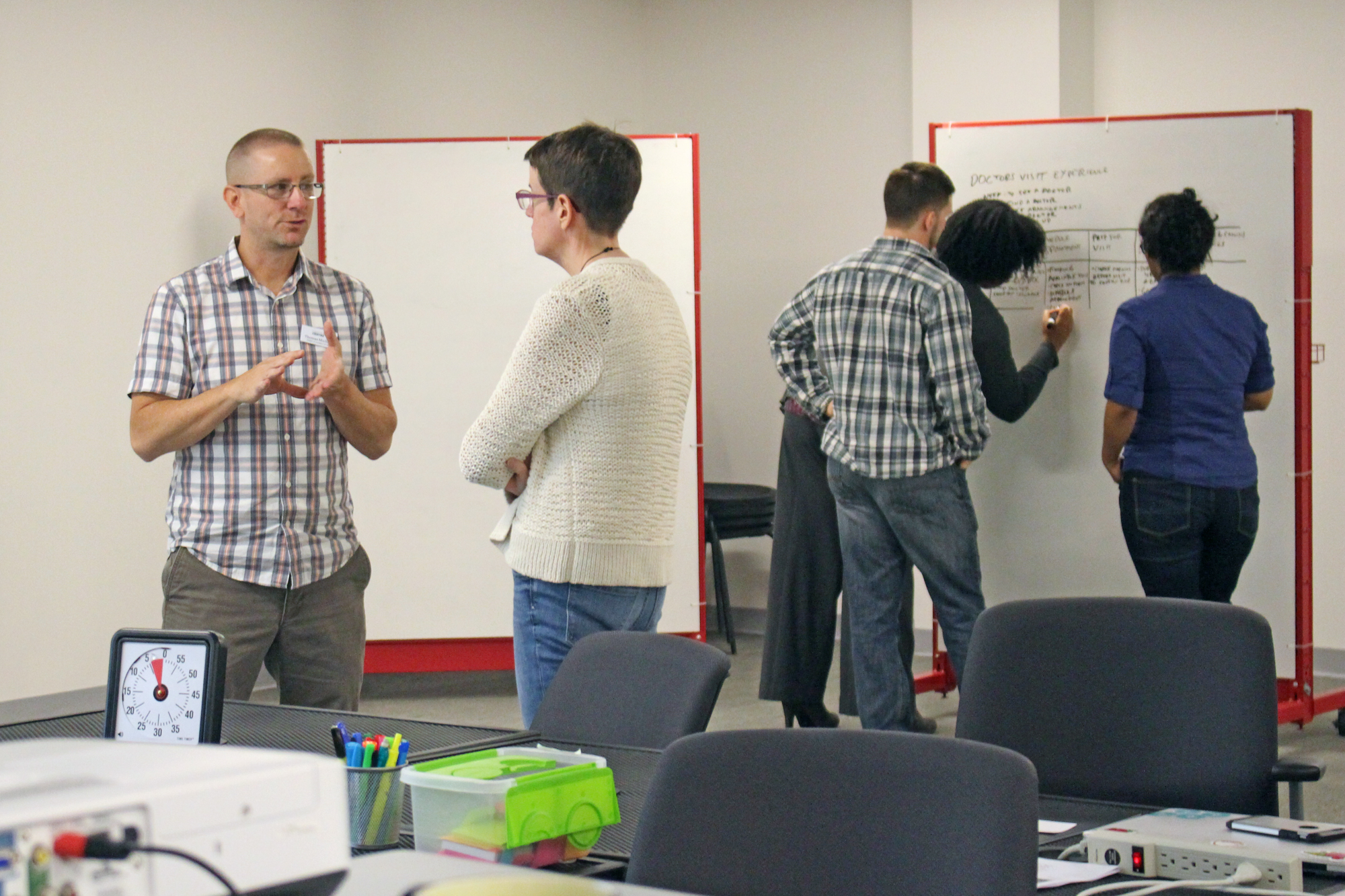 Photo of Facilitator Thomas talking to Kim Goodwin, students working in the background.